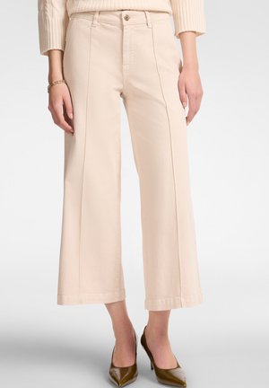Pantalones de pierna ancha en beige, confeccionados en una tela suave, con una cintura alta, costuras verticales y un sutil dobladillo en el bajo. Combinados con tacones puntiagudos.