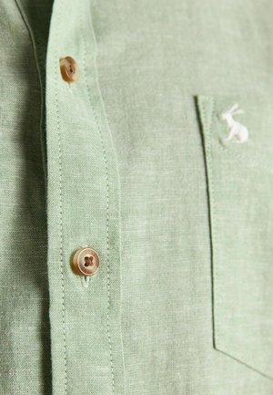 Camicia in lino verde chiaro con bottoni marroni e un piccolo coniglio bianco ricamato sulla tasca sul petto.
