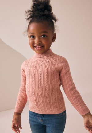Jeune fille souriante aux cheveux bouclés en chignon haut, portant un pull rose torsadé et un jean bleu sur un fond uni.