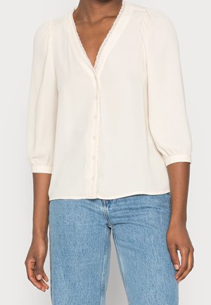 Kvinde iført en cremefarvet V-halsbluse med blonder og knapper, kombineret med lyseblå denimjeans.