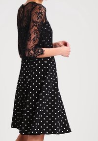 Svart klänning med prickar och utsvängd kjol, med en semi-genomskinlig svart spetsöverdel med ärmar till armbågen och blommönster.