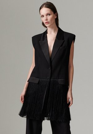 Femme aux cheveux bruns et raides porte un blazer noir sans manches avec des revers en satin et des détails à franges sur un pantalon noir, sur un fond gris.