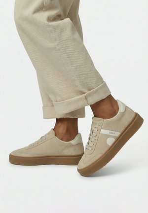 Voeten met beige suède sneakers met gumzolen en opgerolde lichtbeige broeken op een witte achtergrond.