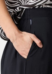 Zwarte broek met een zijzak. Gemaakt van lichte stof, met een gladde textuur en een elastische tailleband. Een label is zichtbaar aan de zijkant.