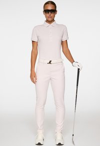 Donna in abbigliamento golfistico rosa pallido, che indossa occhiali da sole e un guanto, tiene un putter da golf mentre si trova di fronte a uno sfondo bianco uniforme.