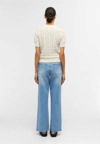 Suéter corto de manga corta en color crema con un patrón de rombos texturizado, combinado con pantalones vaqueros de pierna ancha en azul claro y zapatos de plataforma negros, visto desde la parte de atrás.