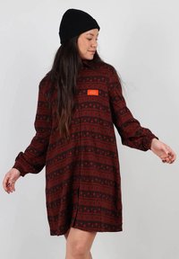 Robe à manches longues de couleur rouge foncé avec un motif comportant des points et des rayures. Comprend un emblème orange proéminent sur la poitrine.