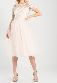 Pale pink tulle dress with a fitted bodice featuring floral lace overlay and short sleeves, knee-length skirt, paired with gold heeled sandals.
