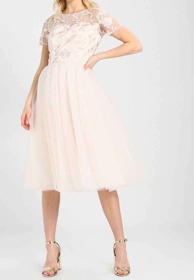 Pale pink tulle dress with a fitted bodice featuring floral lace overlay and short sleeves, knee-length skirt, paired with gold heeled sandals.