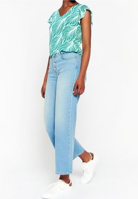 Zielona i biała bluzka we wzór liści z falbanastymi rękawami, zestawiona z jasnoniebieskimi spodniami z wysokim stanem i szerokimi nogawkami oraz białymi trampkami.