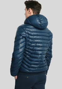 Mężczyzna w ciemnoniebieskiej pikowanej kurtce puchowej z kapturem, stojący z rękami w kieszeniach, odwrócony plecami na szarym tle.