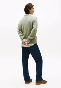 Maglione a maglia verde oliva con colletto e polsini a coste, indossato sopra una camicia a scacchi bianca e verde, abbinato a jeans blu scuro e scarpe marroni.