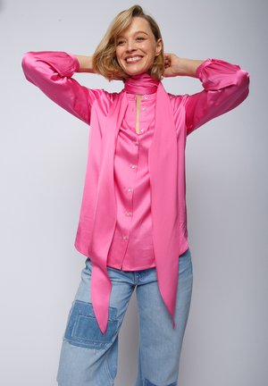 Rosa Satinbluse mit langen Ärmeln, Kragen, Knöpfen und einem passenden Schal um den Hals gebunden, kombiniert mit hellblauen Jeans.