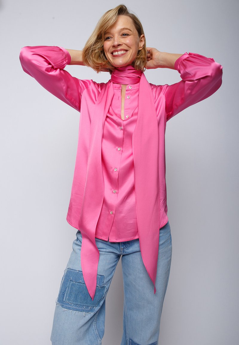 Różowa satynowa bluzka z długimi rękawami, kołnierzykiem, guzikami oraz pasującym szalikiem zawiązanym na szyi, w połączeniu z jasnoniebieskimi dżinsami.
