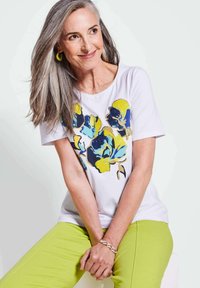 Weißes T-Shirt mit kurzen Ärmeln, das ein buntes Blumendesign in Blau, Gelb und Schwarz zeigt. Das Modell trägt limettengrüne Hosen und ein Armband.