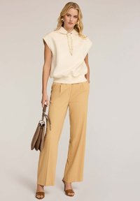 Top sin mangas de color crema con capucha y cordón, combinado con pantalones de pierna ancha de color beige claro. Bolso de cuero marrón y tacones a juego.