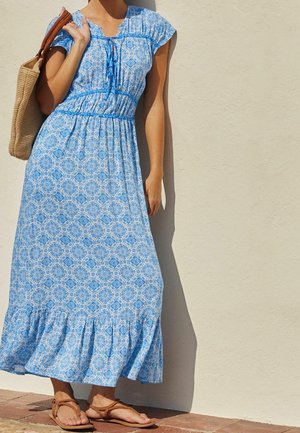 Modrá vzorovaná maxi šaty s krátkými rukávy, nařaseným pasem a rozšířeným lemem. Woven taška a sandály na řemínky doplňují outfit.