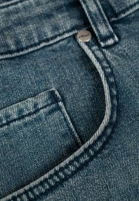 Tela de mezclilla en azul oscuro, con un bolsillo de parche, costuras visibles y un botón metálico con el nombre de la marca grabado.