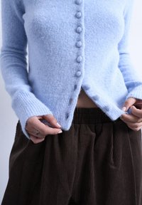 Άτομο που φοράει ανοιχτό μπλε, μαλακό ζακέτα με κουμπιά και σκούρο καφέ παντελόνι κωδρέ, κρατώντας τις άκρες της ζακέτας κοντά στη μέση.