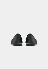 Une paire de ballerines noires avec un petit logo doré sur les côtés externes, présentée de dos sur fond blanc.
