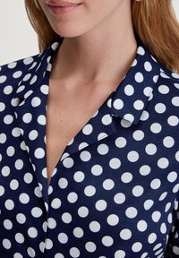 Navyblauwe stoffen blouse met grote witte stippen, voorzien van een notch-kraag en een gladde textuur, weergegeven van de borst omhoog.