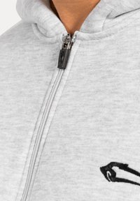 Sudadera con cremallera gris de textura suave, que presenta un logotipo en negro contrastante en el lado izquierdo y una cremallera de metal plateado.