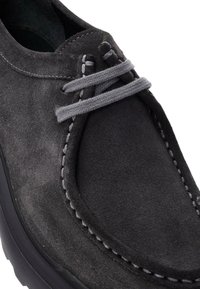 Chaussure en suède gris avec un bout arrondi, dotée de lacets gris et de coutures contrastées. La semelle est noire et légèrement texturée.