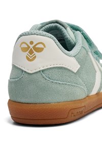 Miętowo-zielony sneaker z cholewką z zamszu, białymi skórzanymi akcentami i brązową gumową podeszwą. Posiada złote logo na pięcie i przeszycia dekoracyjne.