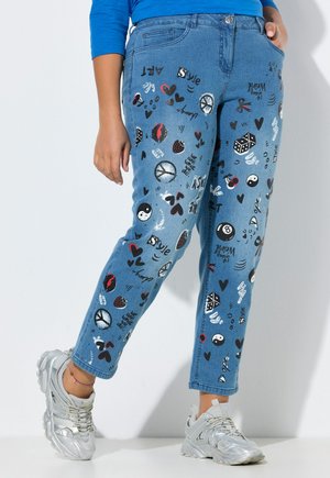 Blå jeans med farverige trykte symboler og ord, båret med sølv sneakers og en blå langærmet top, hænderne afslappet langs siden.