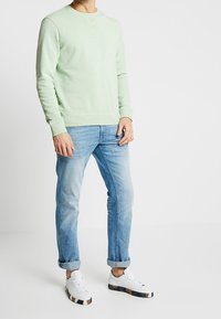 Sudadera verde claro de manga larga, cuello redondo y puños acanalados, combinada con jeans azul claro y zapatillas blancas con suelas con estampado.