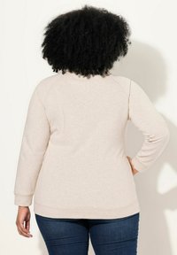 Felpa beige chiaro con una texture morbida, maniche raglan lunghe e polsini a costine. Abbinata a jeans blu scuri. Vista posteriore.
