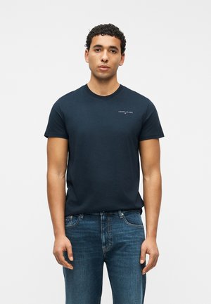 Mladý muž oblečený v tmavomodrom tričku Tommy Jeans a modrých džínsoch, stojaci pred jednoduchým svetlým pozadím.