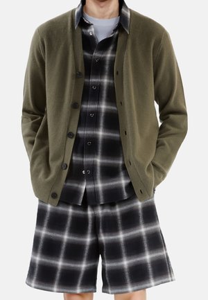Mężczyzna w oliwkowo-zielonym kardiganie zapinanym na guziki, noszący czarno-białą koszulę w kratę i pasujące szorty w kratę, z rękami w kieszeniach, na białym tle.
