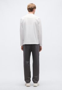 Hosszú ujjú fehér ing kerek nyakkal, szürke nadrággal és világos színű tornacipővel, hátulról ábrázolva, egyszínű háttér előtt.