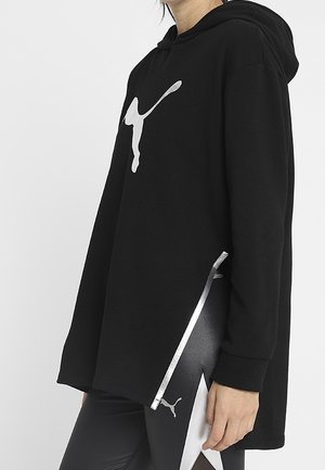 Osoba nosząca czarną bluzę z kapturem Puma z srebrnym logo oraz dopasowane czarne legginsy z srebrnym logo Puma i wzorem po boku.