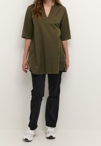 Olivgrünes Tunika mit V-Ausschnitt und kurzen Ärmeln, mit Seitenschlitzen. Kombiniert mit schwarzen Hosen und weißen Sportschuhen.