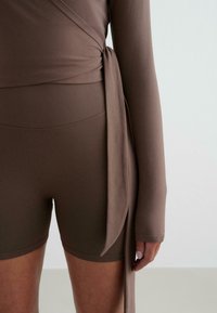 Smeđi wrap top s dugim rukavima, izrađen od mekog materijala. Uparen s odgovarajućim šorcama visoko struka s rebrastom teksturom i bešavnim dizajnom.