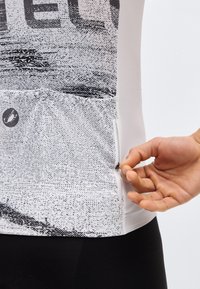 Χέρι ρυθμίζει ένα μικρό φερμουάρ στην πλαϊνή τσέπη ενός λευκού και μαύρου αθλητικού τοπ με στίγματα.