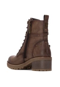 Botín de cuero marrón hasta el tobillo con cremallera lateral, punta redonda y un tacón bajo apilado. Presenta tachuelas decorativas a lo largo de los lados y un upper texturizado.