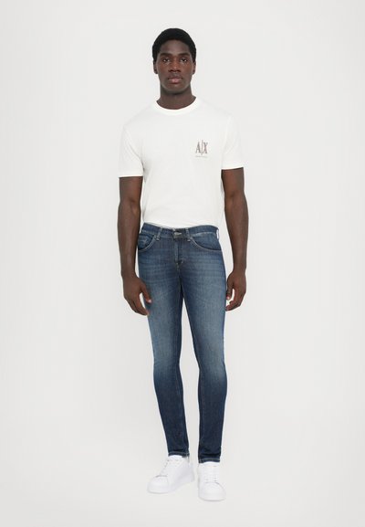 Vit t-shirt med logotyp, mörkblå jeans med lätt blekning, och vita sneakers. Slim-fit design, cottonmaterial, avslappnad stil.