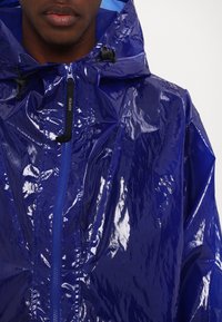 Giacca da pioggia blu brillante con zip frontale e cappuccio regolabile. Realizzata in materiale lucido e impermeabile, presenta dettagli e cuciture nere.