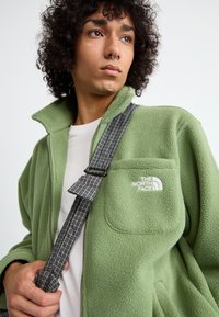 Zelena flis jakna z visokim ovratnikom, zadrgo spredaj in žepi na prsih, ki vsebujejo logo "The North Face". Uporabljena v kombinaciji s karo pasom.