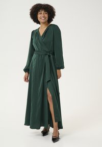 Abito lungo verde avvolgente in tessuto morbido, con maniche lunghe, cintura in vita e uno spacco alto fino alla coscia, abbinato a scarpe nere con punta.