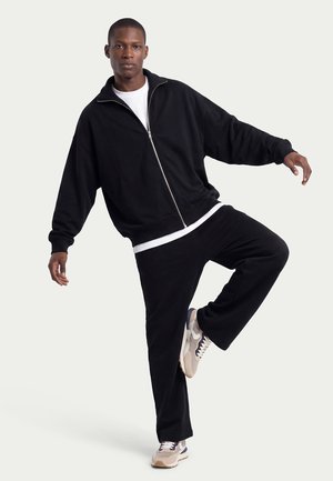 Man balancing on one leg, wearing black zip-up jacket, black pants, white shirt, and beige sneakers, against a plain light background.