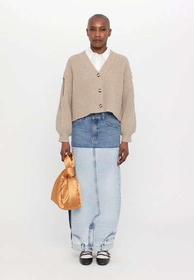 Gilet en tricot beige avec des boutons, chemise blanche à col en dessous, jupe en denim bleu, et un sac en satin doré. Chaussures noires et blanches avec des lanières.