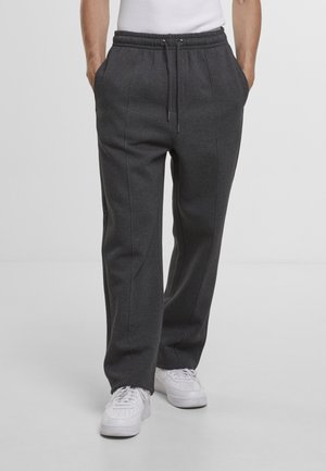 Personne portant un pantalon de survêtement gris foncé à cordon avec des détails de couture, des baskets blanches et un haut blanc sans manches, les mains dans les poches.