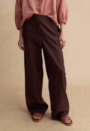 Donna che indossa pantaloni larghi marrone scuro a righe sottili, una camicetta rosa polvere con bottoni e maniche a sbuffo, e sandali aperti con fantasia.