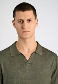 Olivgrön polo-tröja i stickat tyg, med V-hals och korta ärmar. Texturen är mjuk med en subtil lyster.