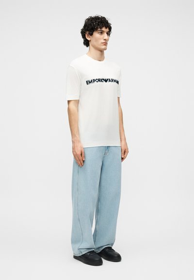 Άνδρας με σγουρά μαλλιά που φοράει λευκό μπλουζάκι Emporio Armani, ανοιχτό μπλε τζιν παντελόνι με φαρδιά μπατζάκια και μαύρα αθλητικά παπούτσια, στέκεται μπροστά σε απλό φόντο.