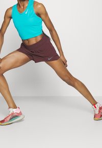 Top corto senza maniche turchese, pantaloni sportivi bordeaux e sneaker multicolori con un logo distintivo e suole texturizzate.